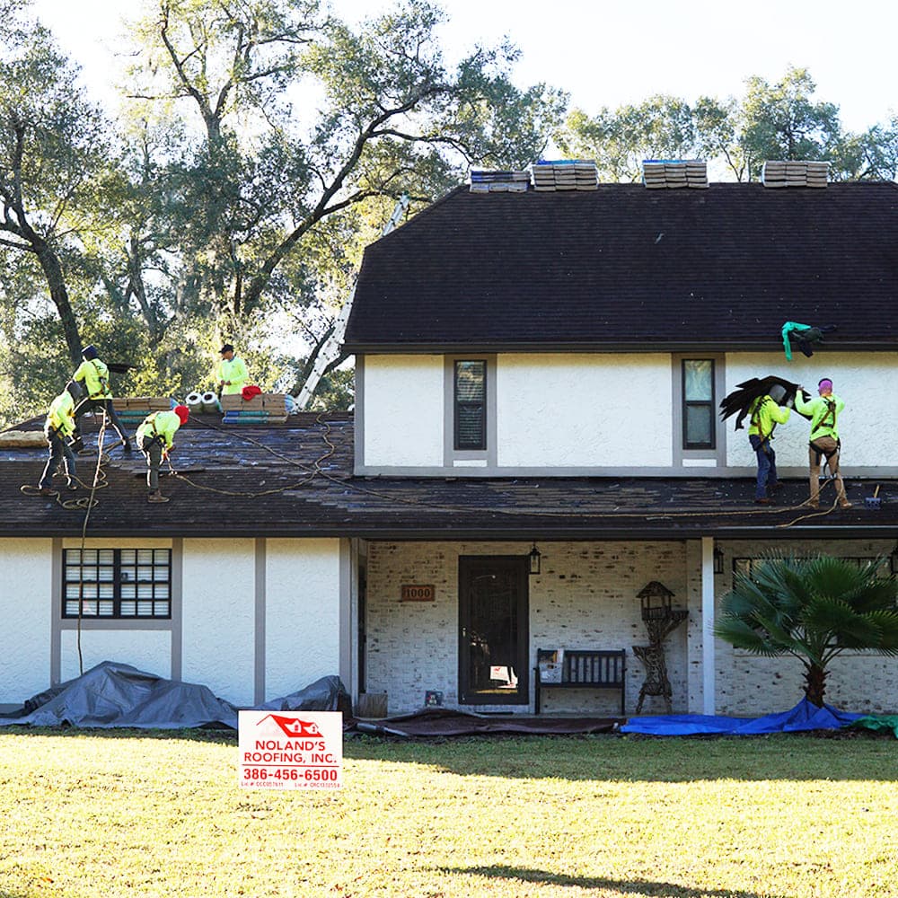 Noland's Roofing crew working on a two-story roof with the company yard sign in the front lawn
