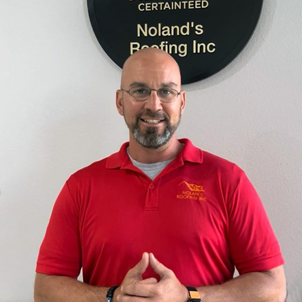 headshot of the Noland's Roofing Branch Manager Ryan taken infront of a white wall with a circular plaque with Noland's Roofing. Ryan is wearing a red branded polo.